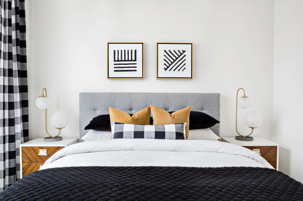 Modern bedroom with black and white buffalo check curtains, geometric artwork, gray upholstered headboard, and gold accent pillows