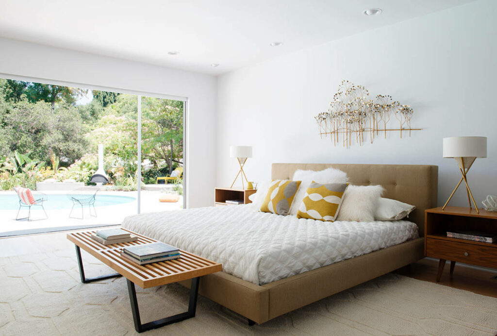 Bright modern bedroom with pool view, tan upholstered bed, wooden bench, and retro-inspired furniture