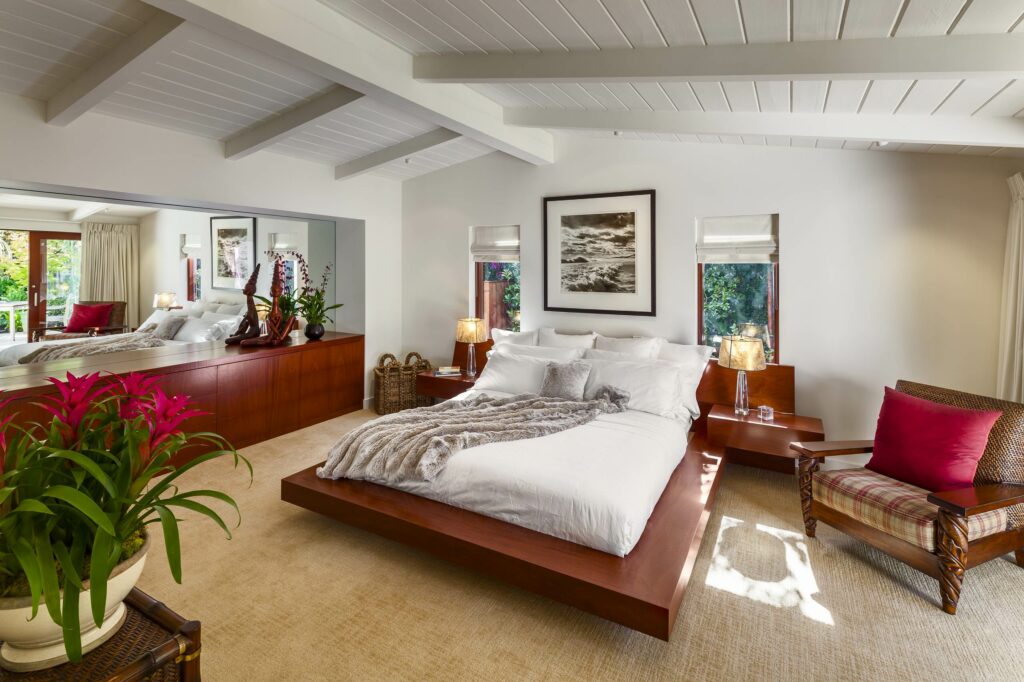 Retro-modern bedroom featuring floating wooden platform bed, vintage rattan chair, exposed beam ceiling, and tropical plants